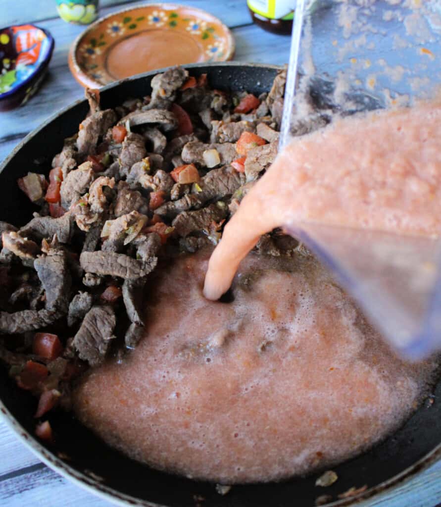 A blender pouring tomato sauce into a skillet with cooked beef.