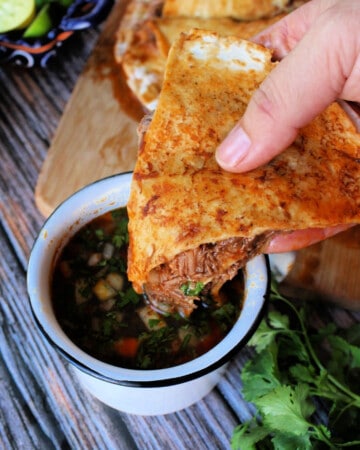 A hand holding a slice of birria pizza and dunking it into a cup with consomme.