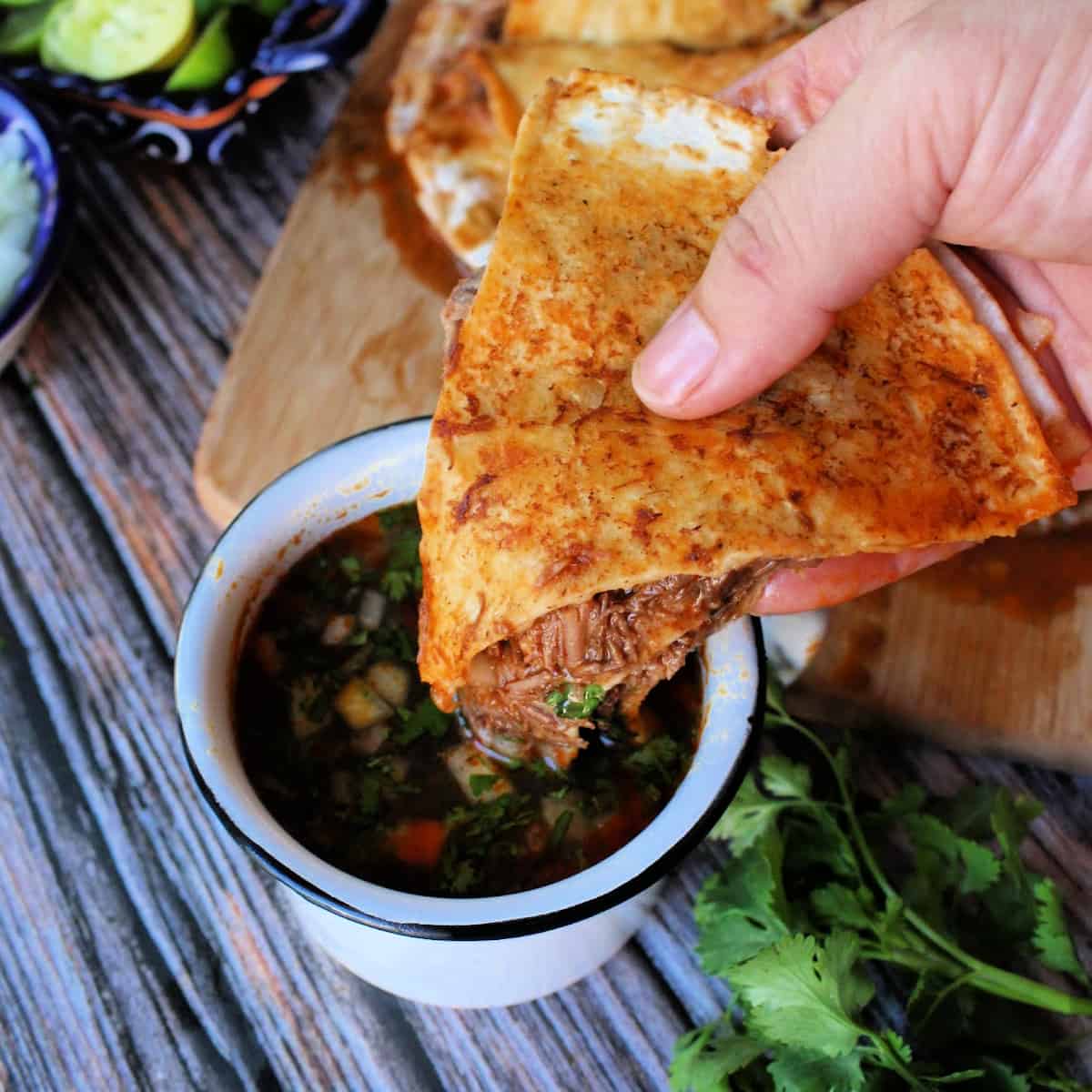 A hand holding a slice of birria pizza and dunking it into a cup with consomme.