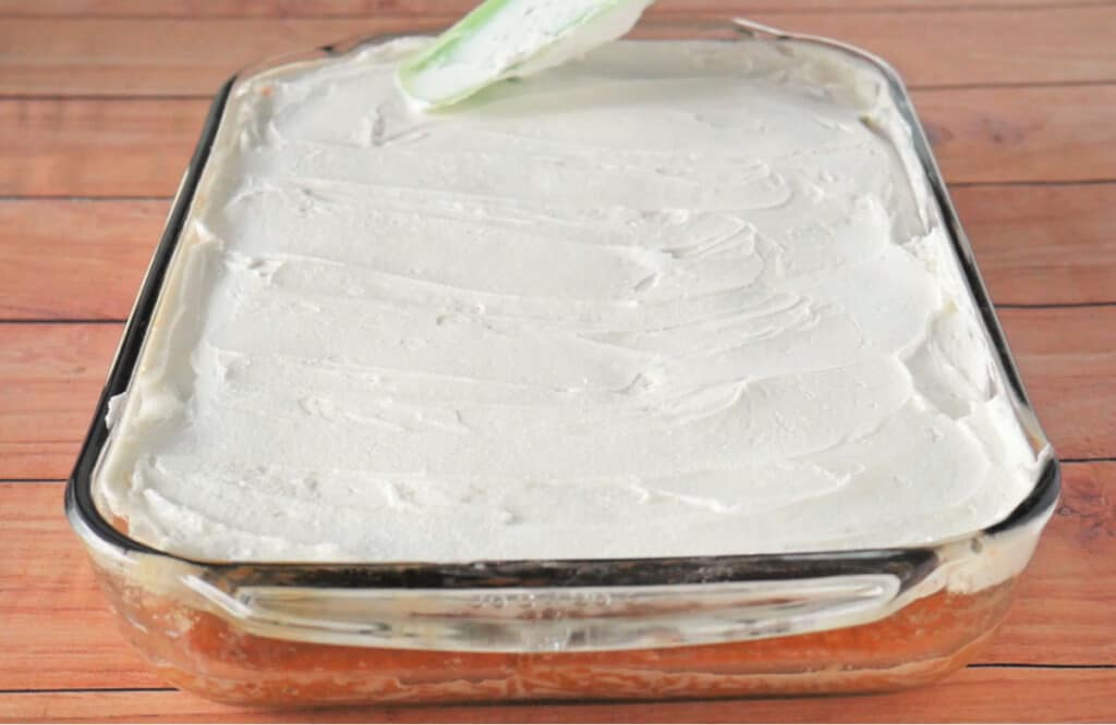 Whipped cream being spread all over a cake in a glass baking dish.