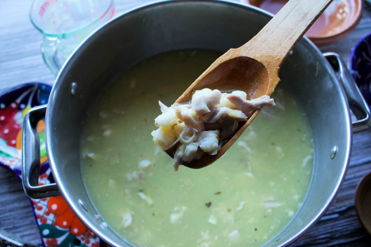 A wooden spoon with green pozole over a cooking pot.