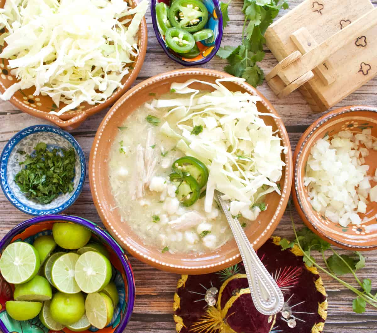 A picture of green chicken pozole served in a Mexican clay plate surrounded by all the toppings.