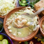 A spoon with pozole verde over the bowl and the toppings.