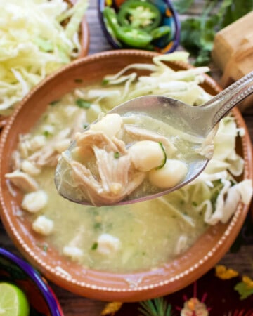 A spoon with pozole verde over the bowl and the toppings.