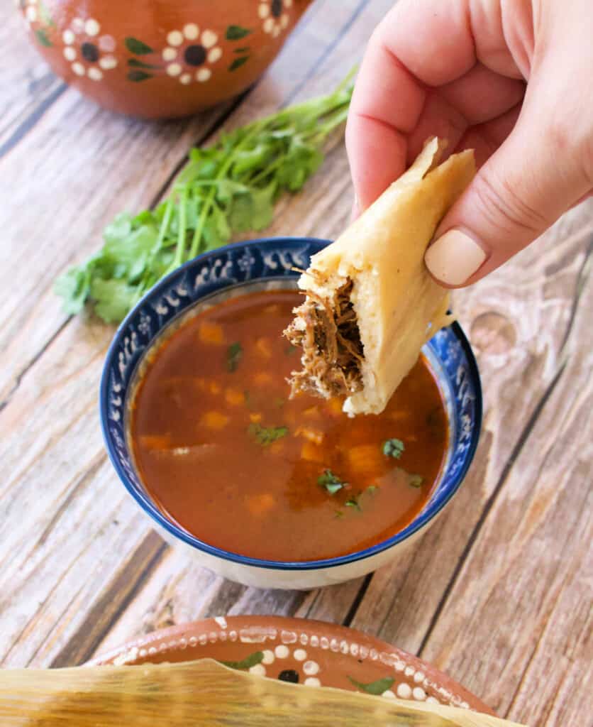 A hand dunking Beef Birria Tamales into a bowl with consomme.