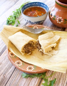 A birria tamal sitting on a corn husk cut in half and sitting next to the consomme.