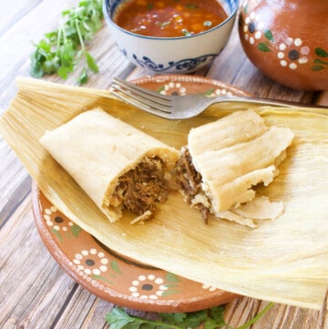 A birria tamal sitting on a corn husk cut in half and sitting next to the consomme.