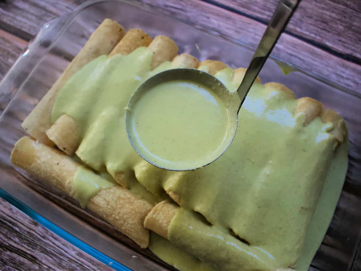 A ladle with creamy green sauce over unbaked enchiladas.