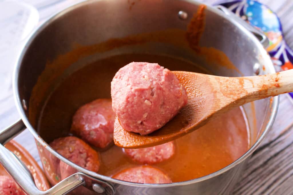 A wooden spoon holding a raw meatball over a stock pot.