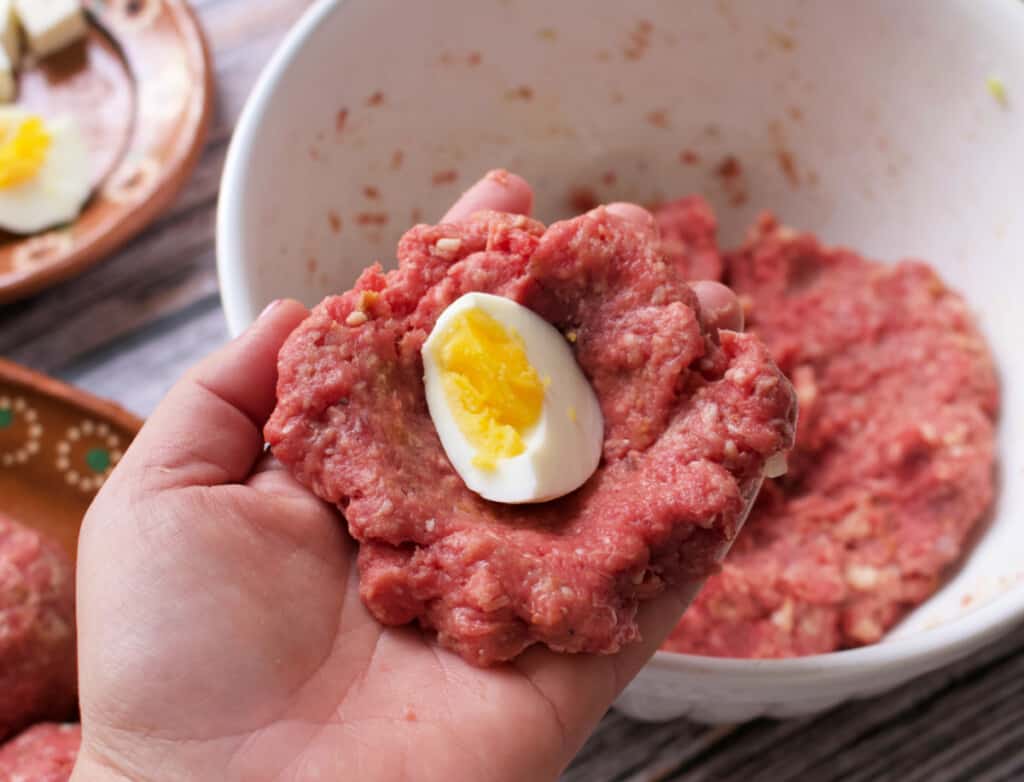 A hand holding flattened ground beef with an egg in the center.
