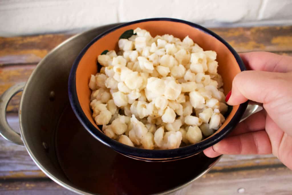 A hand holding a bowl full of hominy above a pot.
