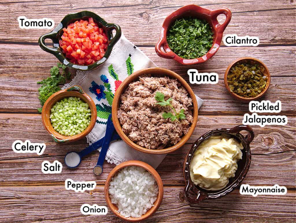The ingredients needed to make Mexican Tuna Salad laid out and labeled on a wooden surface.