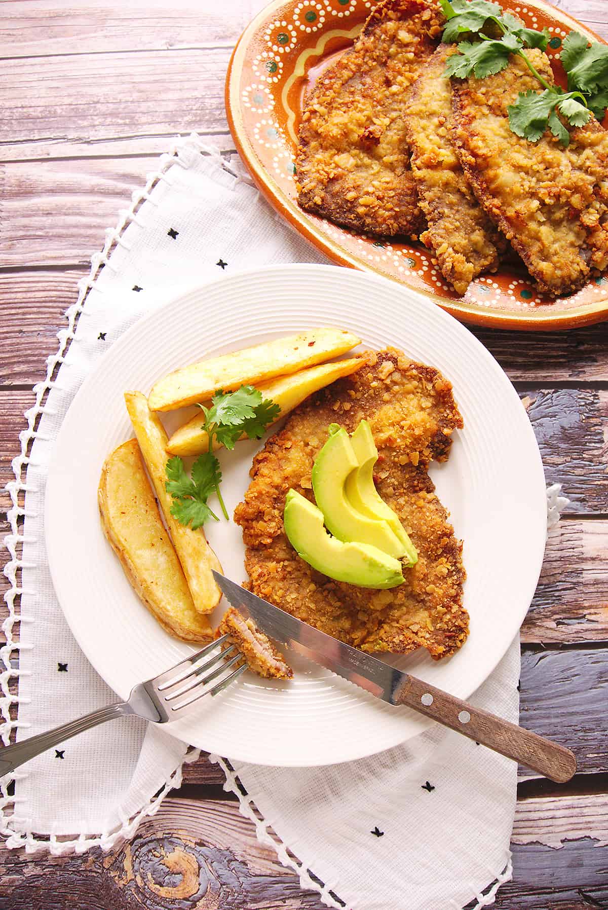 Milanesa de Res served with avocado slices on top and potato fries on a glass plate