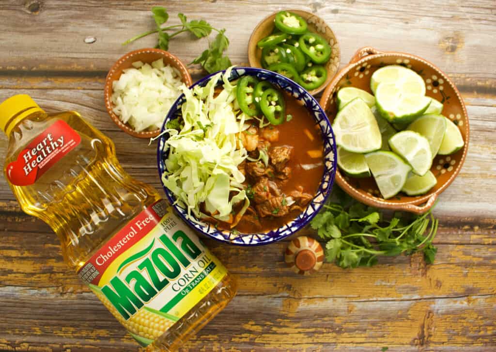 A bowl of red beef pozole topped with cabbage and surrounded by more toppings and a bottle of Mazola oil.