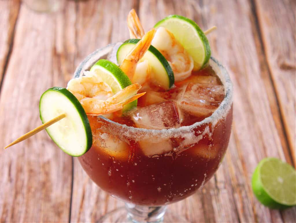 Michelada de Camaron served in an ice cold glass with a salted rim and a skewer of shrimp and cucumbers.