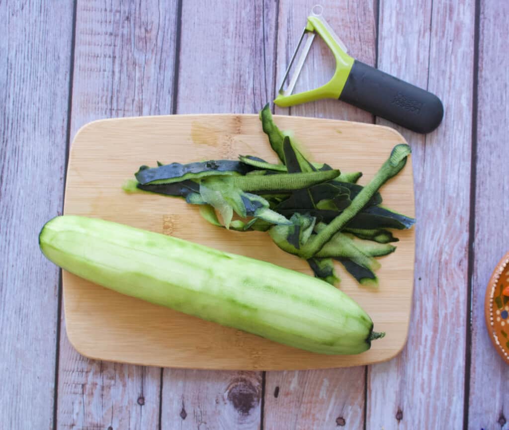 A picture of a peeled cucumber next to a peeler and the shaved peel.