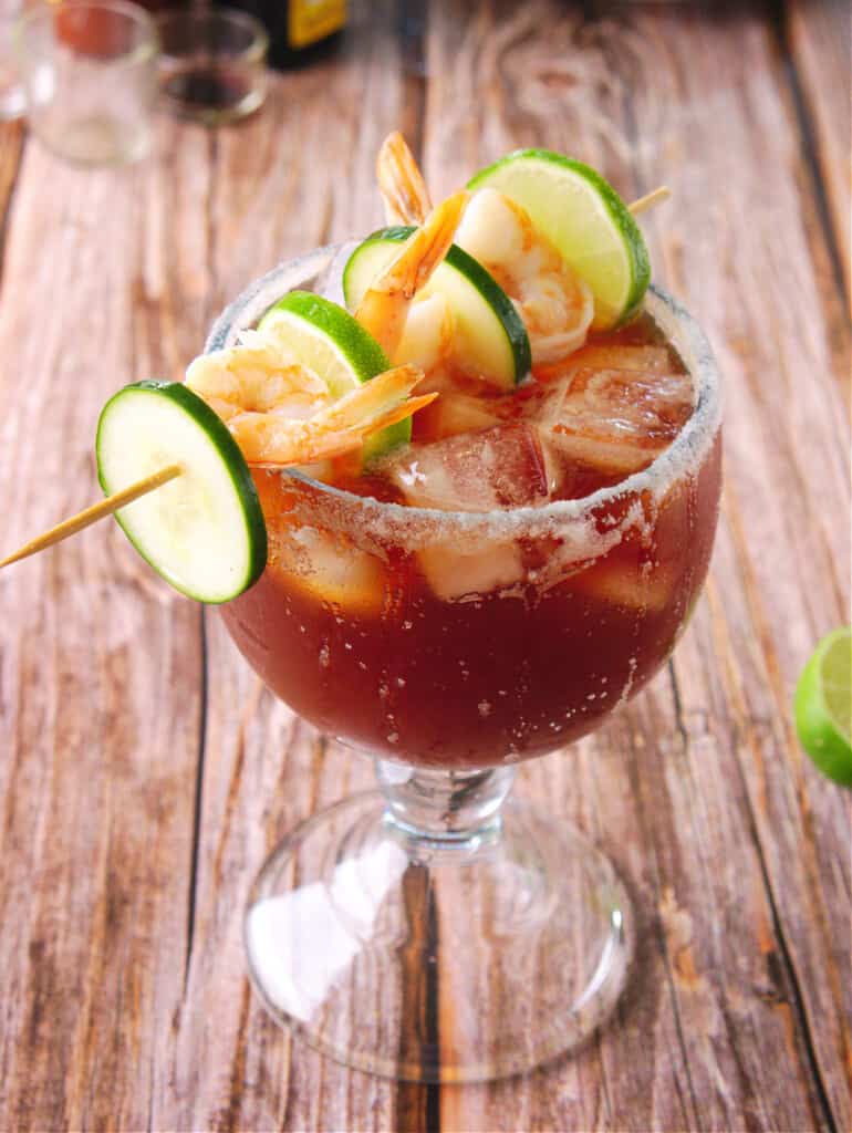 A shrimp michelada served in an ice cold glass and topped with a skewer of shrimp and cucumbers.