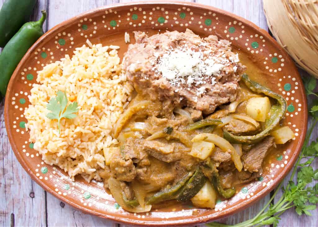 Bistec Ranchero served on a decorative Mexican clay plate next to beans and rice.