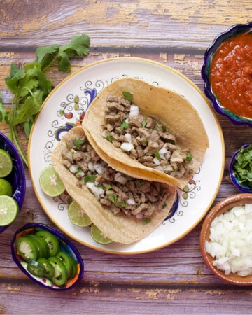 Two tacos de bistec served on a plate and surrounded by the toppings.