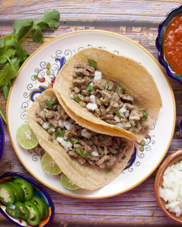 Two tacos de bistec served on a plate and surrounded by the toppings.