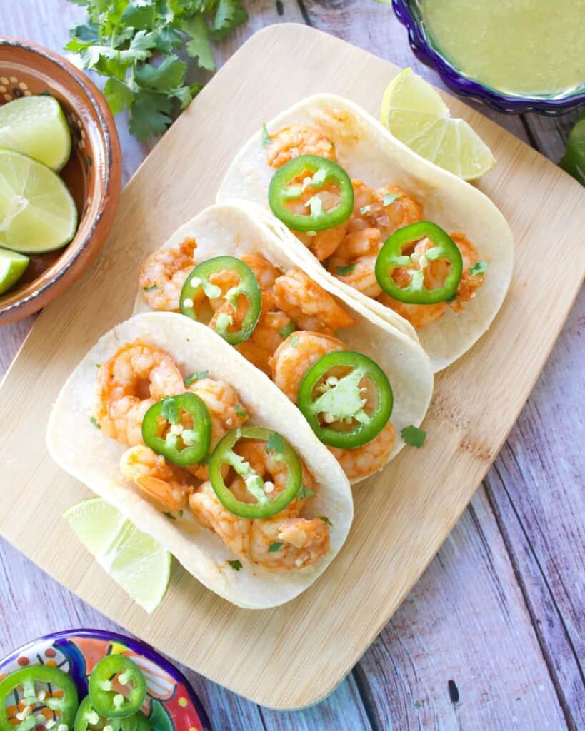 Three shrimp tacos topped with jalapeno slices sitting on a wooden cutting board.