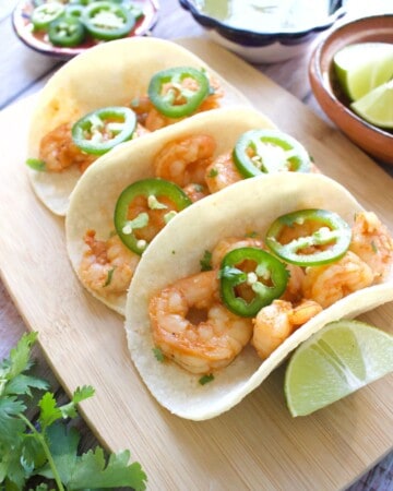 Three shrimp tacos topped with jalapeno slices sitting on a wooden cutting board.
