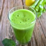 A glass of bright green jugo verde sits on a wooden surface next to celery stalks and a spinach leaf.
