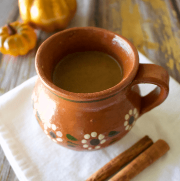 Pumpkin Champurrado served in a decorative Mexican clay mug next to pumpkins and cinnamon sticks.