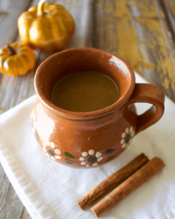 Pumpkin Champurrado served in a decorative Mexican clay mug next to pumpkins and cinnamon sticks.