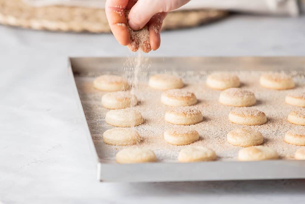 A hand sprinkling sugar and cinnamon on freshly baked Pan de Polvo cookies.