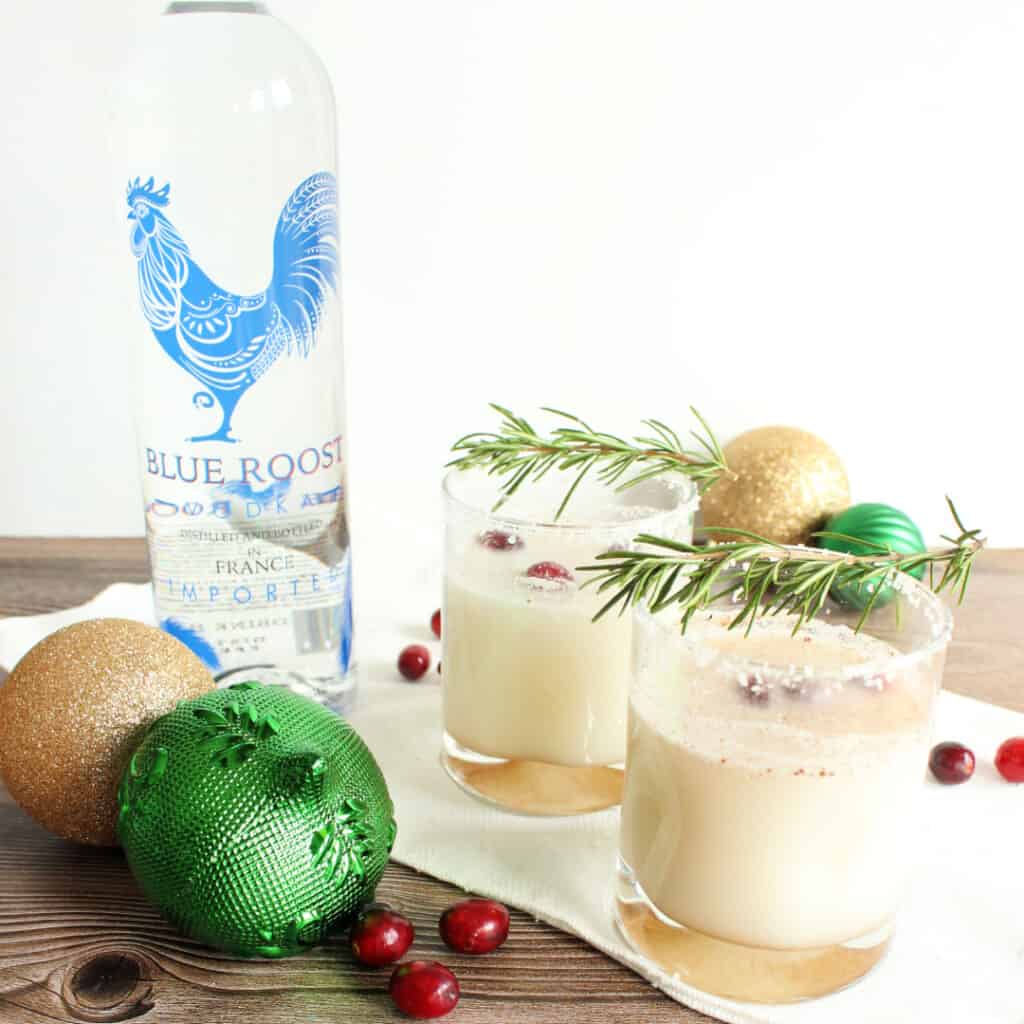 Two glasses of Coconut Christmas Coconut topped with sprigs of rosemary and surrounded by tree ornament, cranberries, and vodka bottle.