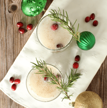 Two glasses of Coconut Christmas Coconut topped with sprigs of rosemary and surrounded by festive tree ornament and cranberries.