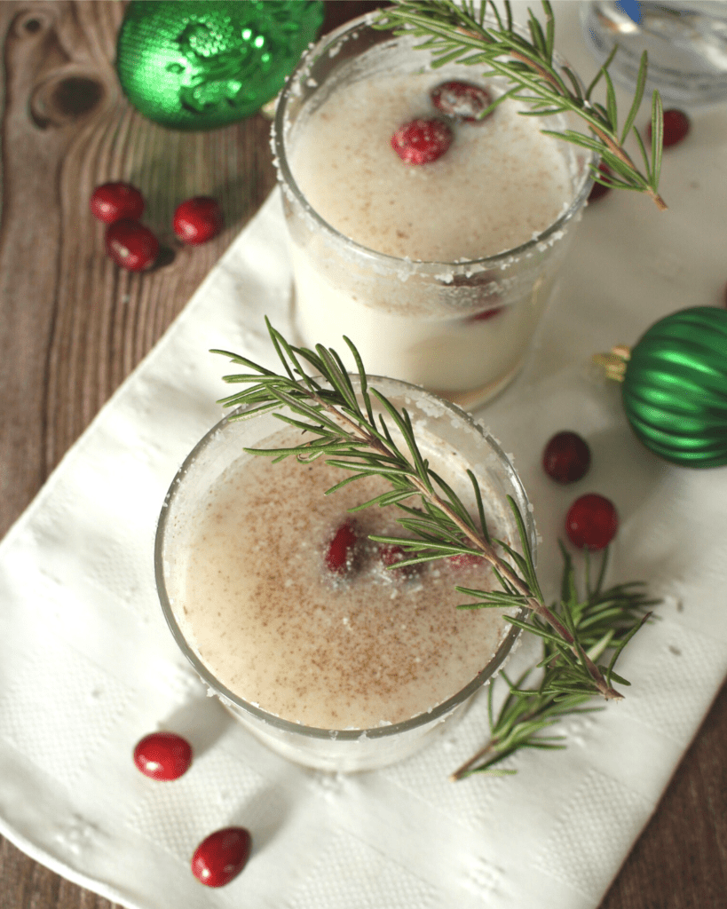Two glasses of Coconut Christmas Coconut topped with sprigs of rosemary and surrounded by festive tree ornament and cranberries.