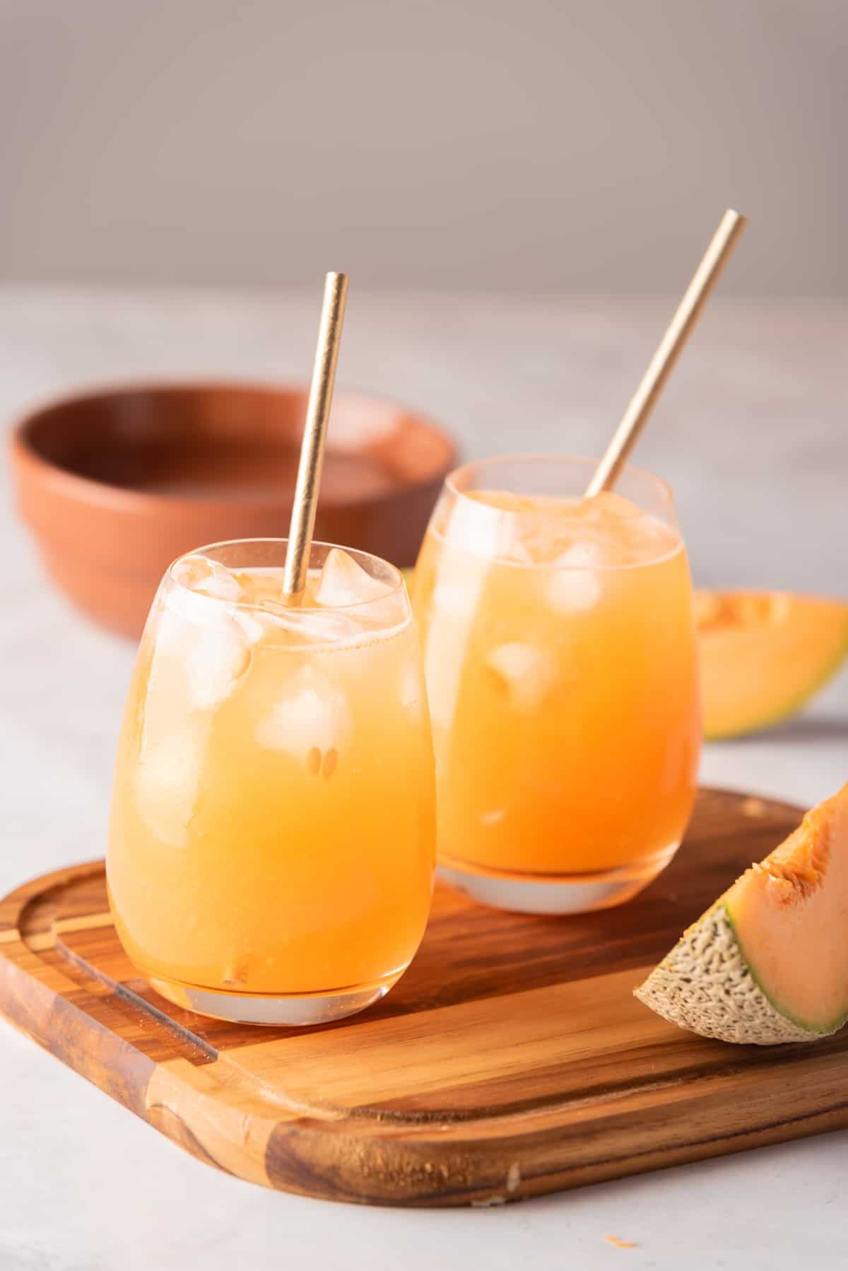 Two glasses served with Cantaloupe Agua Fresca sitting on a wooden cutting board.