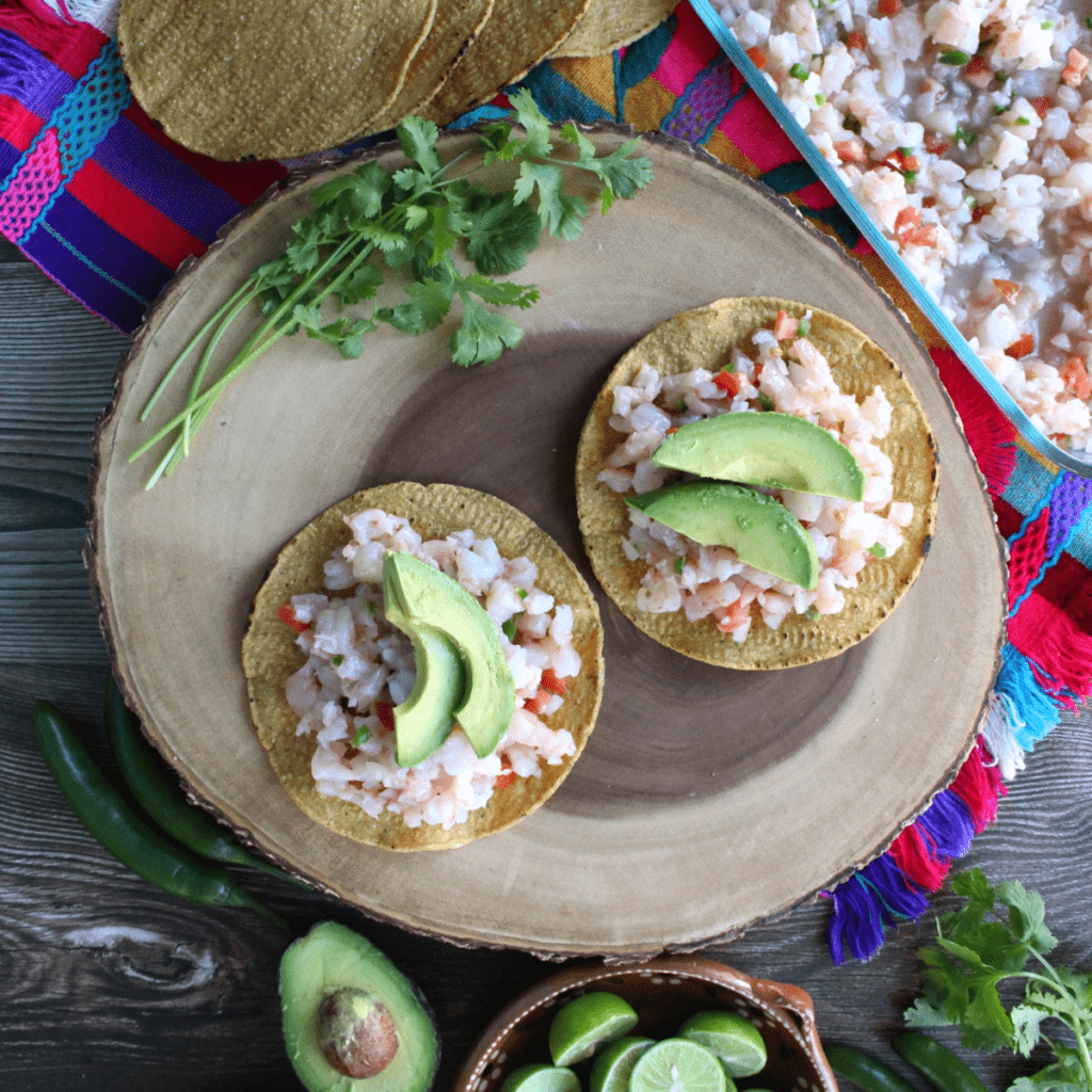 Two tostadas of Shrimp Ceviche topped with sliced avocado sitting on top of a wooden surface.