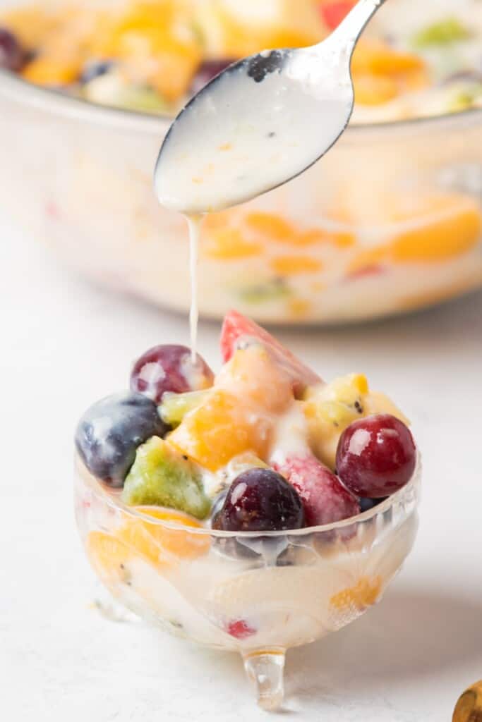 A spoon drizzling the creamy sauce over the fruit salad.