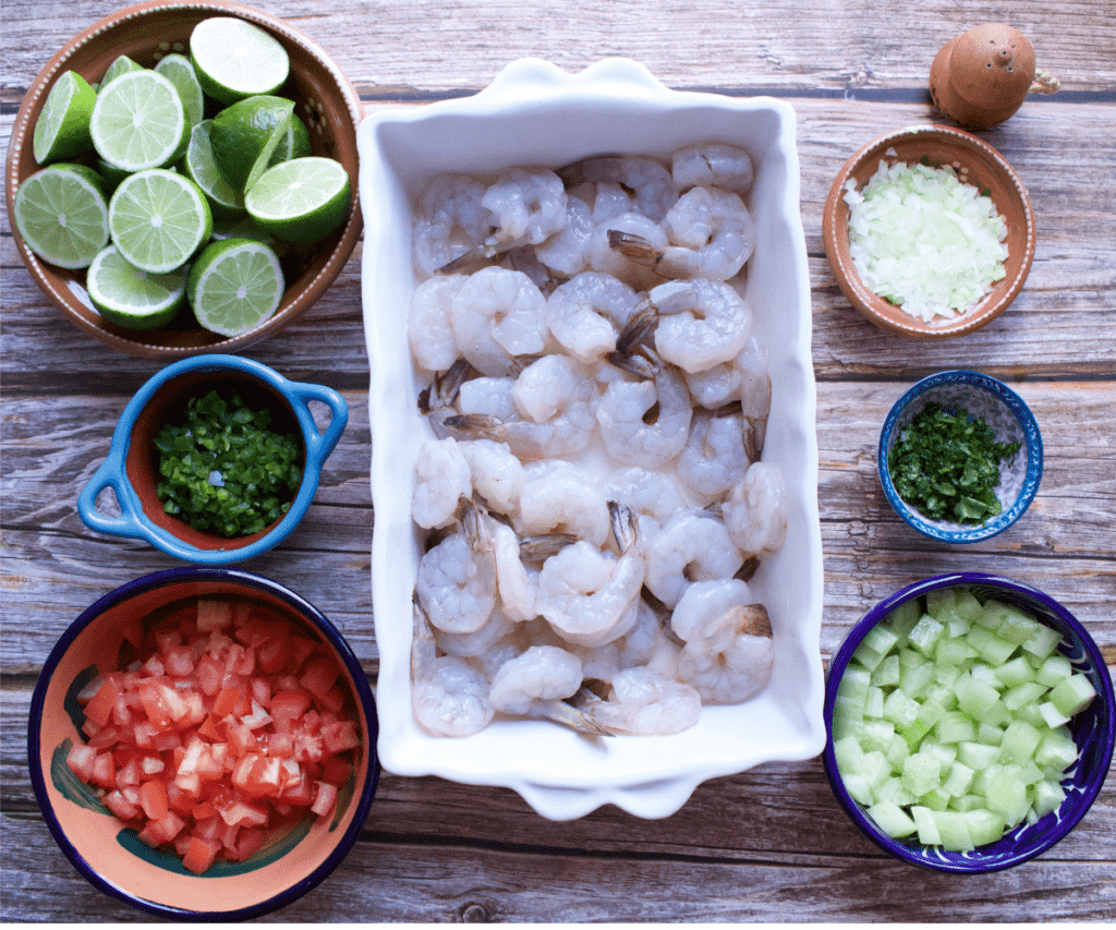 Raw shrimp and the rest of the ingredients needed to make ceviche de camaron sitting on a wooden table.