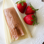 An unwrapped strawberry tamal sitting on a white plate next to three fresh strawberries.