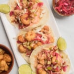 Baja Shrimp Tacos served on a white next to lime wedges and pickled red onion.