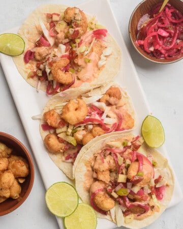 Baja Shrimp Tacos served on a white next to lime wedges and pickled red onion.