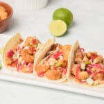 Three Baja Shrimp Tacos served on a white next to lime wedges.