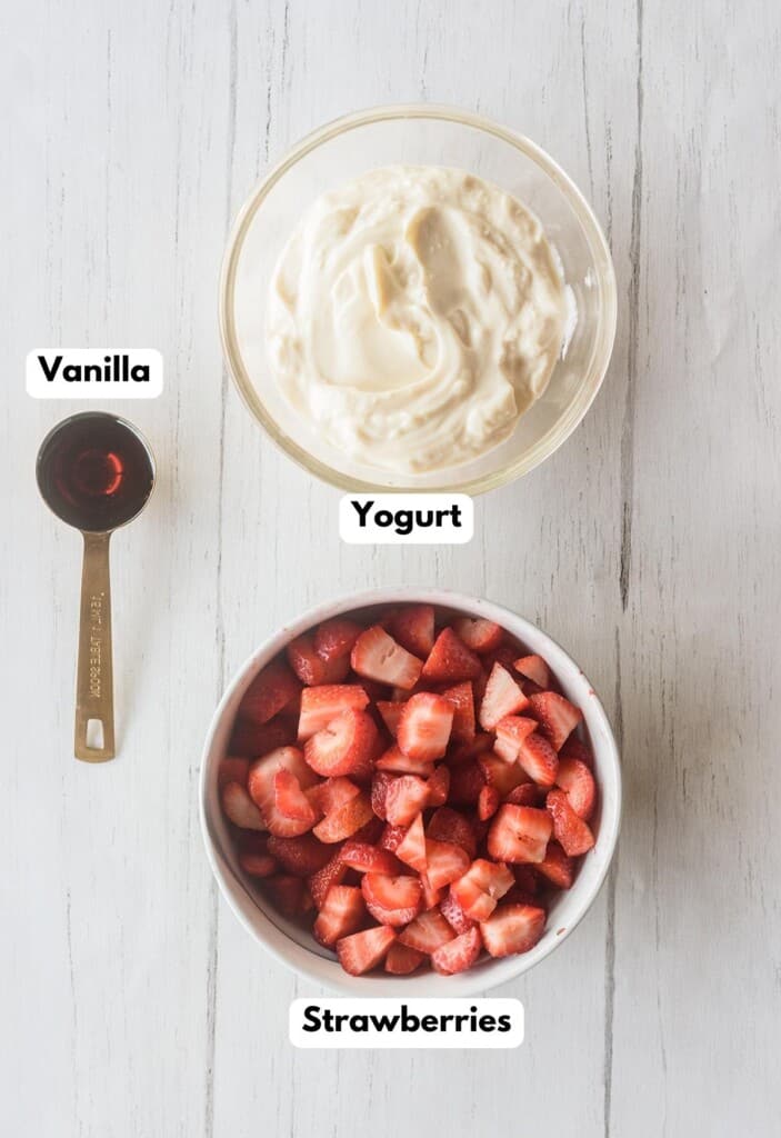 The ingredients needed to make Strawberry Yogurt Popsicles labeled and sitting on a wooden surface.
