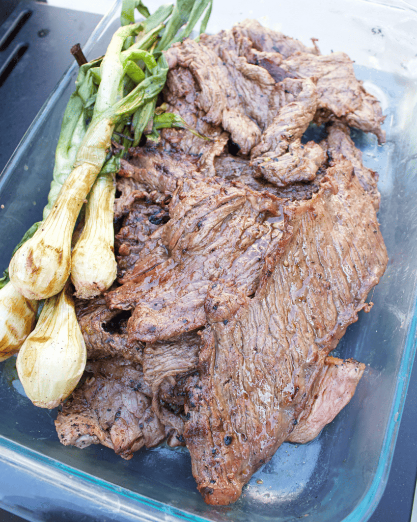 Grilled carne asada sitting on a glass baking dish next to cambray onions.