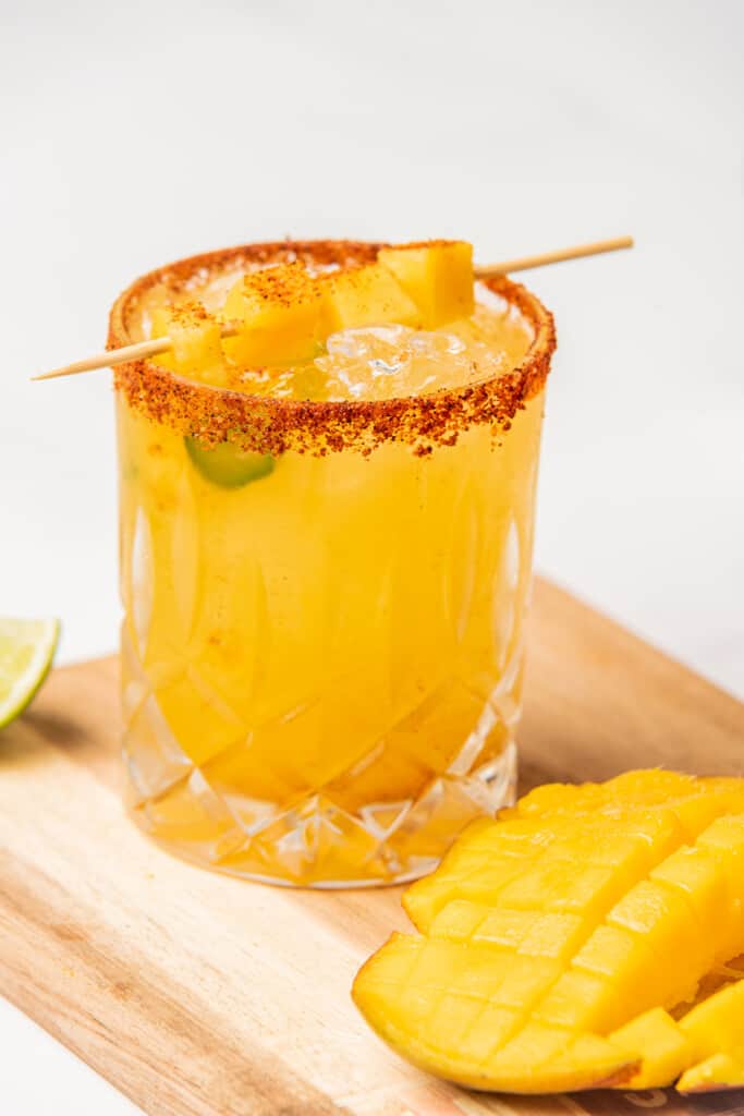 A Spicy Mango Margarita served in a glass with a rim of Tajin and a skewer of mango pieces.