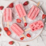 Strawberry Yogurt Popsicles sitting on a ice and surrounded by fresh strawberries.
