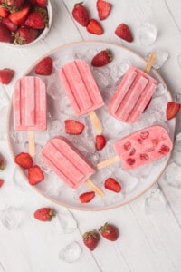 Strawberry Yogurt Popsicles sitting on a ice and surrounded by fresh strawberries.