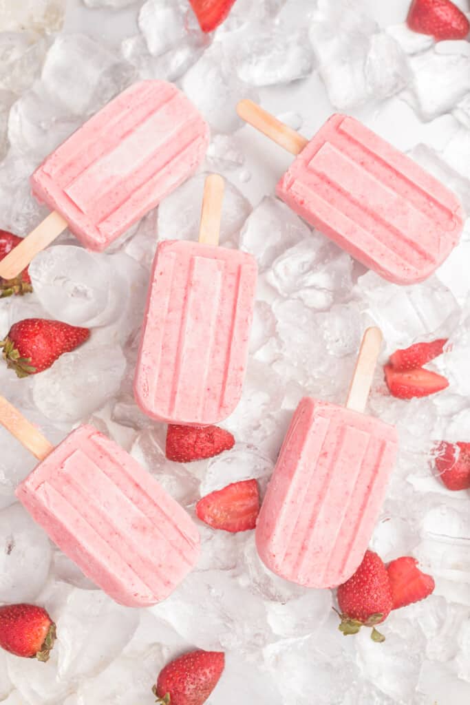 Strawberry Yogurt Popsicles sitting on ice cubes and surrounded by fresh strawberries.
