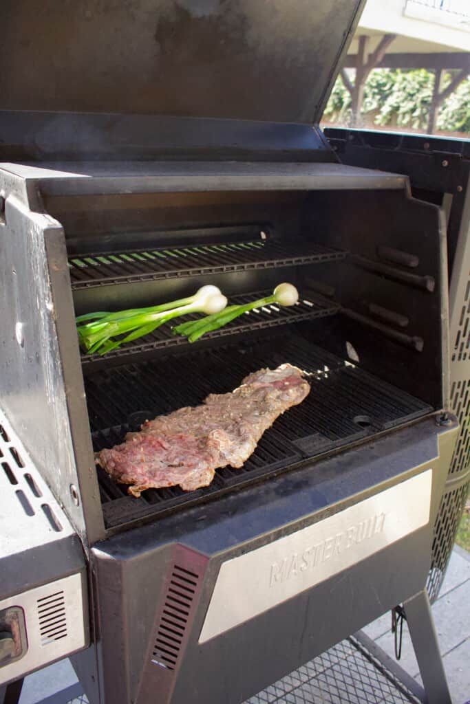 Flap steak cooking on an outdoor grill with green onions cooking on the upper level of the grill.