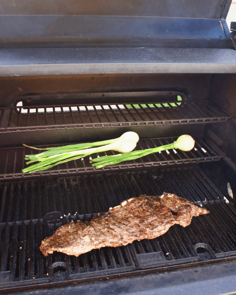 Flap steak grilling on an outdoor grill with green onions cooking on the upper grill level.