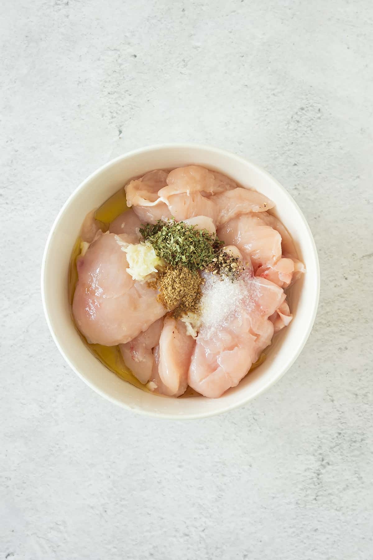 Raw chicken marinating in a white bowl with spices.
